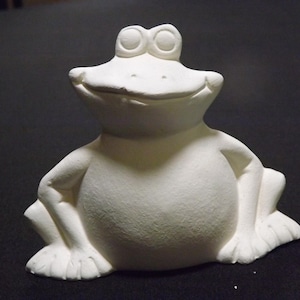 Könnte beinhalten: Eine weiße Keramik-Froschfigur mit einem lächelnden Gesicht. Der Frosch hat große Augen, einen runden Körper und die Arme liegen an den Seiten an. Die Figur ist unbemalt und bereit zur individuellen Gestaltung.