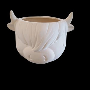 May include: A white ceramic planter shaped like a Highland cow's head. The planter features detailed sculpting of the cow's face, including horns, fur, and a small nose. The interior is a light tan color, and the planter is set against a black background.