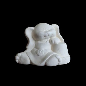 Puede incluir: Figura de cerámica blanca de un conejito sentado sosteniendo un pequeño barril. El conejito tiene orejas grandes, una cara redonda y lleva un babero con un diseño de estrella. La figura no está pintada y está sobre un fondo negro.