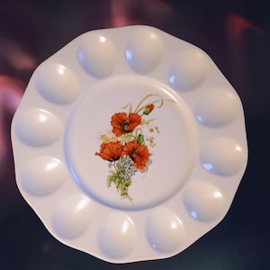 May include: White ceramic paint palette with a scalloped edge and 12 wells. The center features a floral design with red poppies and blue wildflowers. The palette is round with a glossy finish.