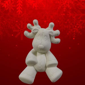 Puede incluir: Figura de reno de cerámica blanca con diseño cosido, sobre fondo rojo con motivos de copos de nieve. El reno tiene grandes cuernos, una nariz pequeña y ojos de botón, ideal para la decoración navideña.