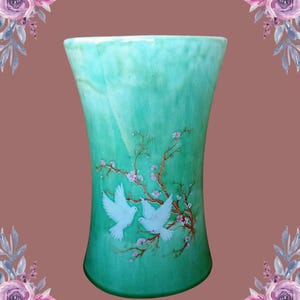 May include: A turquoise ceramic vase with a flared top. The vase features a painted design of two white doves perched on a flowering branch. The vase is a decorative item.