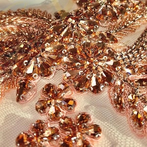May include: Close-up of a delicate, rose gold beaded fabric with intricate floral patterns. The fabric is adorned with sparkling rhinestones and beads, creating a luxurious and elegant look.