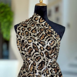 Könnte beinhalten: Ein One-Shoulder-Kleidungsstück auf einer schwarzen Schaufensterpuppe. Der Stoff ist mit goldenen und schwarzen Pailletten in einem Leopardenmuster bedeckt. Das Kleidungsstück ist drapiert, um das Design und die Textur zu zeigen.