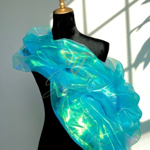 May include: A vibrant, iridescent blue fabric draped over a black dress form. The fabric is gathered and ruffled, creating a voluminous, flowing effect. The lighting highlights the fabric's shimmering quality, suggesting a formal or artistic garment.