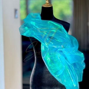 May include: A turquoise iridescent fabric draped over a black dress form. The fabric has a flowing, ethereal quality, with hints of green and blue. The dress form has a gold-coloured neck and is set against a blurred background.