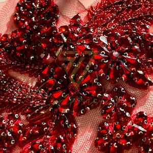 May include: Red beaded applique with a floral design. The applique is made of small, round beads and sequins. It is perfect for adding a touch of sparkle to any project.