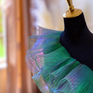 Puede incluir: Un maniquí negro con cuello y cabeza dorados. El maniquí muestra una sección de una prenda con volantes superpuestos, verdes y morados iridiscentes. Los volantes tienen una textura plisada y crean un efecto dramático.