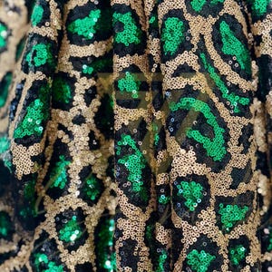 Op de afbeelding: Close-up van een stof met een luipaardprint. De stof heeft een patroon van zwarte en groene pailletten op een gouden achtergrond. De pailletten zijn dicht op elkaar gepakt, waardoor een glinsterend effect ontstaat.