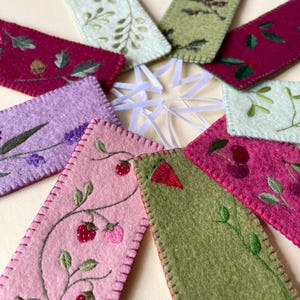 May include: A selection of handmade felt bookmarks in shades of pink, green, and purple. Each bookmark is embroidered with botanical designs like strawberries, lavender, and leaves. The edges are stitched with a contrasting thread.