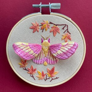 May include: Embroidered moth and fall leaves in a wooden hoop. The moth has pink and yellow wings, with a yellow body. The leaves are in shades of red, orange, and yellow. The background is a beige fabric.