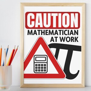 May include: A framed print with the text "CAUTION MATHEMATICIAN AT WORK." The sign features a red and white warning symbol with a calculator, a black Pi symbol, and a light wood frame. A glass with colored pencils is in the foreground.