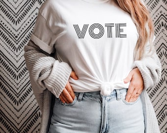 May include: A white t-shirt with the word "VOTE" printed in black, with multiple lines for each letter. The shirt is worn by a person with long brown hair, and is tied at the waist.