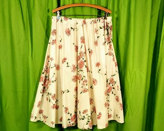 May include: A cream-colored skirt with a floral pattern of pink flowers and green stems. The skirt is hanging on a wooden hanger against a green curtain backdrop. The skirt has an elastic waistband.