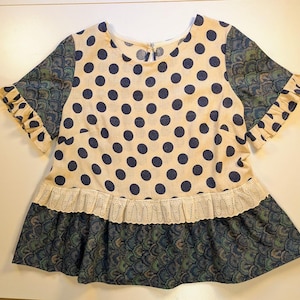 May include: A cream-colored blouse with a navy blue polka dot pattern. The blouse has a ruffled neckline and short sleeves with a blue and green floral pattern. The bottom of the blouse has a white lace trim and a blue and green floral skirt.