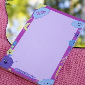 May include: A pink notepad with a purple border and a light purple interior. The border is decorated with cartoon planets, stars, and clouds. The top of the notepad has a blue cloud with the word "Note:" written in white.