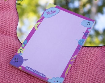 May include: A pink notepad with a purple border and a light purple interior. The border is decorated with cartoon planets, stars, and clouds. The top of the notepad has a blue cloud with the word "Note:" written in white.