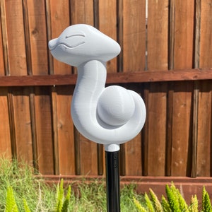 May include: A white, stylized garden stake featuring a smiling, serpentine design. The sculpture is mounted on a black metal pole. The background includes a wooden fence and green foliage.