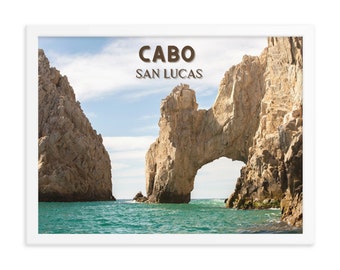 May include: A framed photograph of a natural rock arch formation in the ocean. The arch is made of light brown rock and is surrounded by blue water. The text "CABO SAN LUCAS" is printed in brown letters at the top of the image.