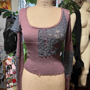 May include: A mauve long-sleeved top with a scoop neckline and patchwork design. The sleeves and upper chest feature grey, distressed fabric. The top is displayed on a Wolf Model Form Co. mannequin.