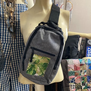 May include: A gray crossbody bag with a black strap and a floral patch pocket. The bag has a zipper closure and a small pocket on the front.
