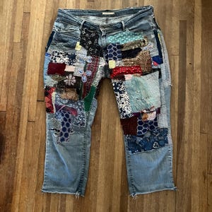 Op de afbeelding: Lichtblauwe spijkerbroek met patchwork details. Het patchwork heeft verschillende stoffen in tinten blauw, rood en wit, met zichtbare stiksels. De jeans heeft een klassiek ontwerp met vijf zakken en een rafelige zoom.