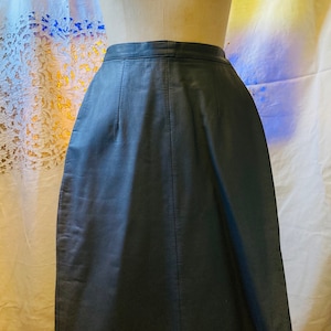 May include: A dark gray leather skirt with a straight, A-line silhouette. The skirt has a simple design with a center seam and a zipper closure.