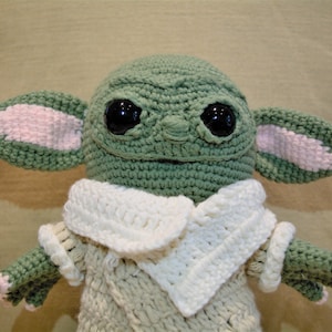 May include: A crocheted plush toy resembling a character from the Star Wars franchise. The toy is green with large, black eyes and pink ears. It is wearing a white crocheted collar and a beige crocheted sweater.