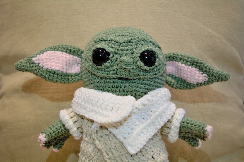 May include: A crocheted plush toy resembling a character from the Star Wars franchise. The toy is green with large, black eyes and pink ears. It is wearing a white crocheted collar and a beige crocheted sweater.