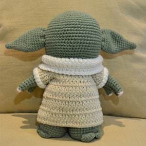 May include: A crocheted green and beige plush toy with large ears and a white collar. The toy is wearing a beige crocheted dress.