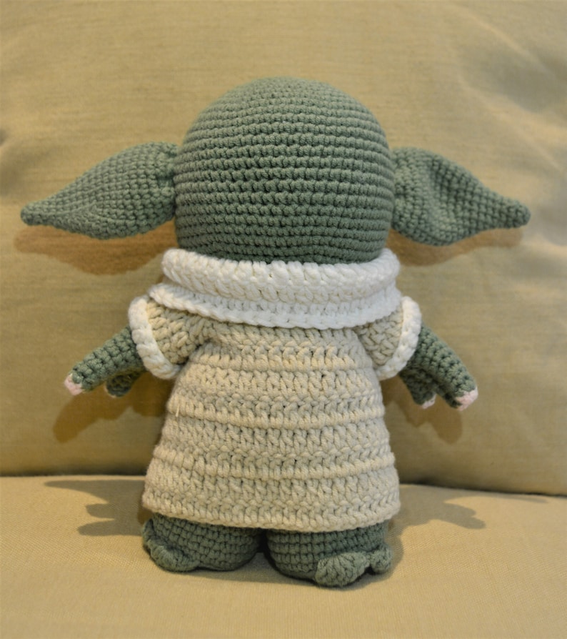 May include: A crocheted green and beige plush toy with large ears and a white collar. The toy is wearing a beige crocheted dress.