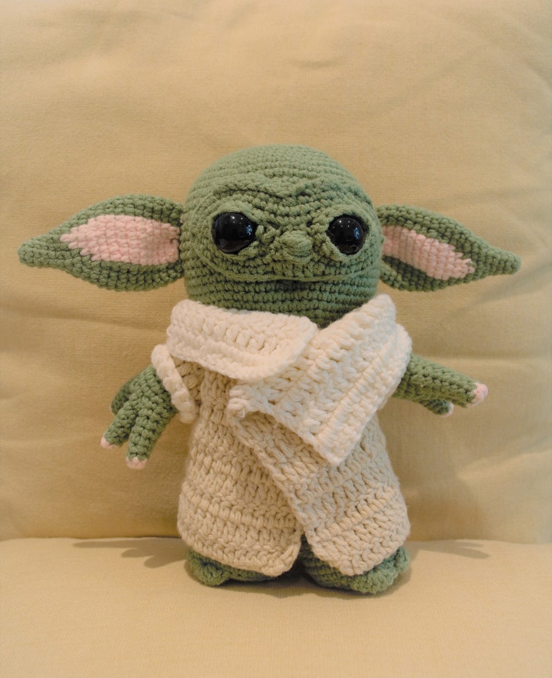 May include: A green crocheted plush toy with large black eyes and pink ears, wearing a white crocheted coat.