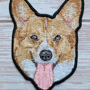 以下が含まれることがあります： コーギー犬の顔が刺繍されたパッチ。パッチは黒い縁取りで、茶色、白、ピンクの色合いで詳細なステッチが施されています。犬の舌はピンク色で、鼻は黒色です。パッチは、生地にアイロンをかけたり、縫い付けたりするように設計されています。