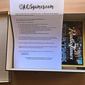 May include: A white sheet of paper with instructions on how to assemble a game board. The paper is in a brown cardboard box with a game board inside. The game board is a black and white image of the Star Trek logo with the words "Radio Times Official Collector's Edition" above it.