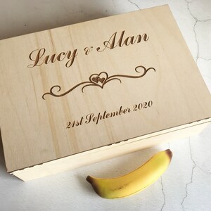 May include: A wooden box with the inscription "Lucy & Alan 21st September 2020" and a decorative heart design. The box is light brown and has a natural wood finish.