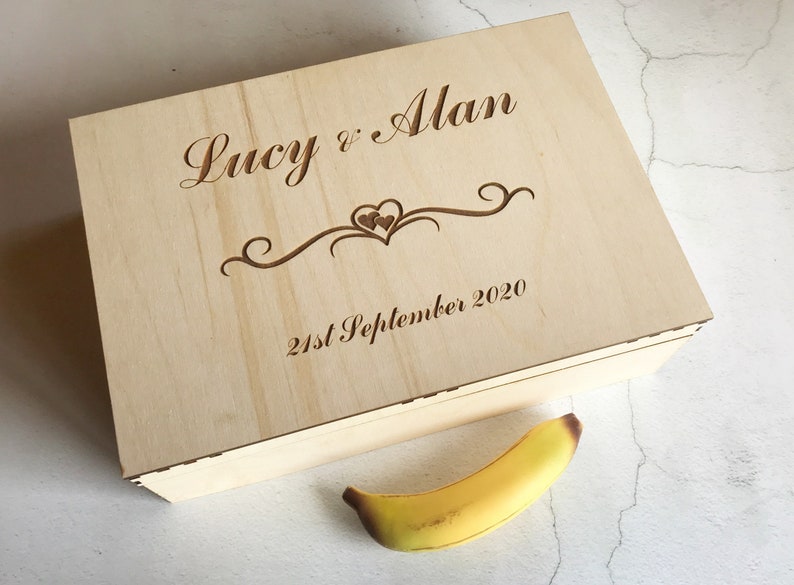 May include: A wooden box with the inscription "Lucy & Alan 21st September 2020" and a decorative heart design. The box is light brown and has a natural wood finish.