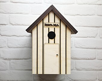 May include: A wooden birdhouse shaped like a beach hut with a brown roof and white siding. The birdhouse has a small round entrance hole and a rectangular door. The text "Beach Hut" is written on the front of the birdhouse.