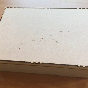 May include: A plain, light brown wooden box with a dark brown border around the top and bottom edges. The box has a simple, rectangular shape.