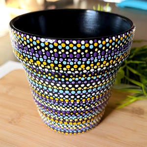 May include: A black ceramic flower pot with a colorful dot pattern. The dots are in shades of purple, yellow, blue, and white. The pot has a wide opening and sits on a wooden surface. The interior of the pot is black.