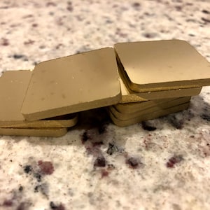 Könnte beinhalten: Ein Stapel von acht quadratischen Untersetzern in Goldfarbe. Die Untersetzer sind aus einem glatten, harten Material gefertigt und haben einen leicht erhöhten Rand.