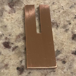 May include: A copper-colored metal U-shaped object. The object has a rectangular base with two parallel, upright sections forming the U shape. The surface has visible brush marks, indicating a brushed metal finish. The object is likely a tool or a decorative item.