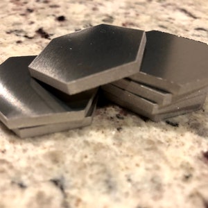 May include: A stack of six silver hexagonal coasters. The coasters are smooth and have a slightly rounded edge.