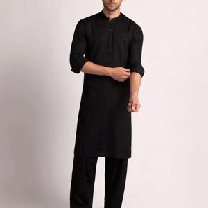 May include: Black kurta pajama set. The outfit features a long tunic with a mandarin collar and button-down front, paired with loose-fitting trousers. The sleeves are rolled up. The ensemble is completed with black shoes. The clothing is made of a solid black fabric.