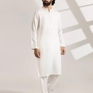 May include: A white, long-sleeved tunic and matching pants set. The tunic has a mandarin collar and a buttoned placket. The pants are straight-legged. The outfit is made of a lightweight fabric. The person is wearing brown leather shoes.