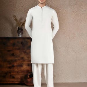 May include: A man wearing a white, long-sleeved tunic and matching trousers. The tunic has a mandarin collar and a buttoned placket. The outfit is a traditional style, suitable for formal or cultural events. The background is a neutral, warm tone.