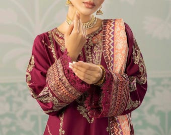 May include: A burgundy embroidered dress with intricate gold detailing. The dress features long sleeves with ornate cuffs and a matching scarf with a geometric pattern. The model is wearing gold jewelry, including earrings and a necklace.