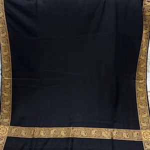 May include: A black shawl with a decorative gold border featuring a paisley design. The shawl has a fringe along the bottom edge. The gold border adds a touch of elegance to the dark fabric.