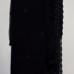 May include: A black sheer fabric shawl featuring a wide, intricate lace border with subtle sequin embellishments. The main body of the shawl is adorned with small, scattered floral-shaped embroidery details in matching black thread.