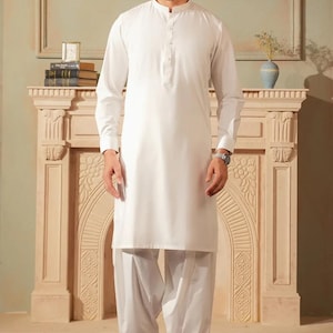 May include: A white, long-sleeved tunic and salwar kameez outfit. The tunic has a mandarin collar and buttoned placket. The salwar, or loose trousers, are gathered at the ankles. The outfit is worn with black leather shoes. The background features a decorative fireplace.