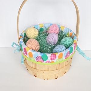 May include: A woven basket with a wooden handle filled with pastel colored crocheted Easter eggs. The basket is lined with a colorful fabric with a polka dot pattern.
